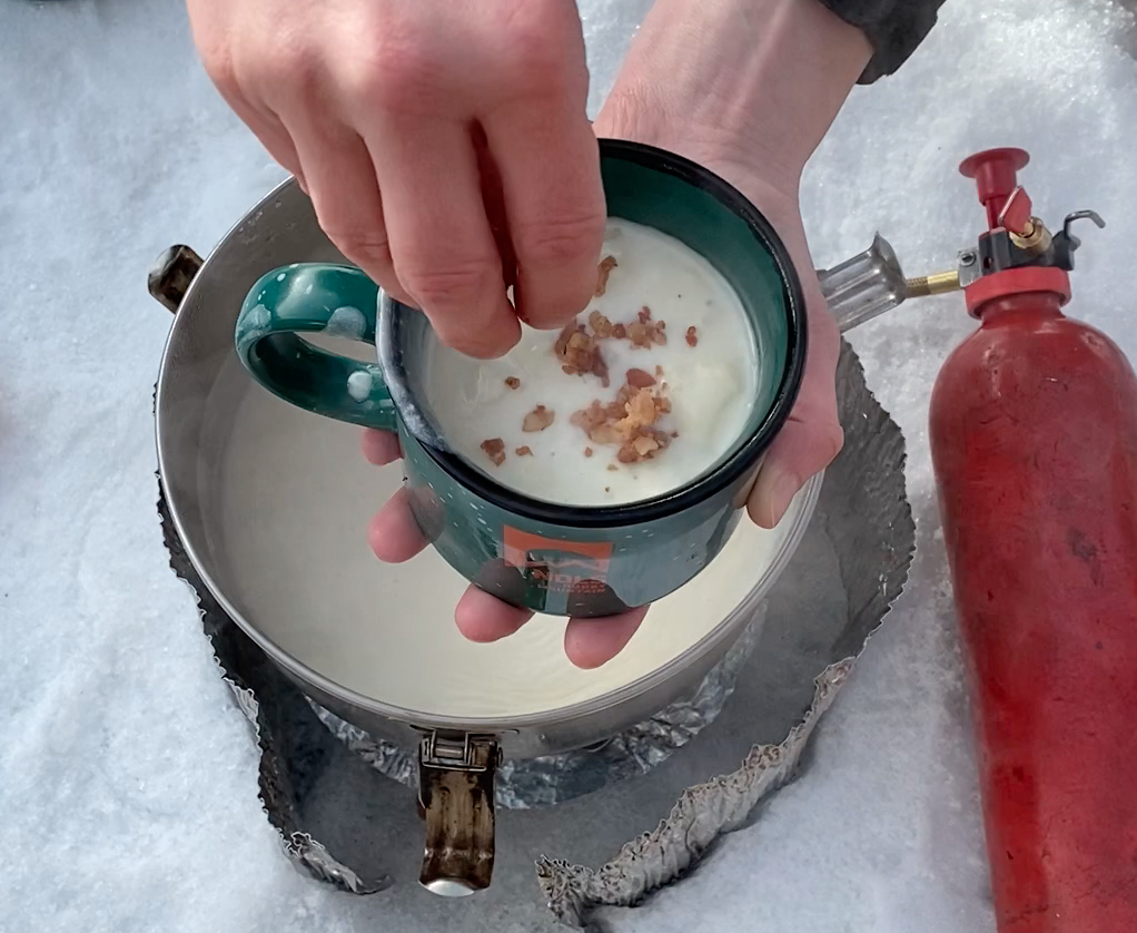 hands sprinkle bacon bits onto potato soup in a mug over a WhisperLite stove