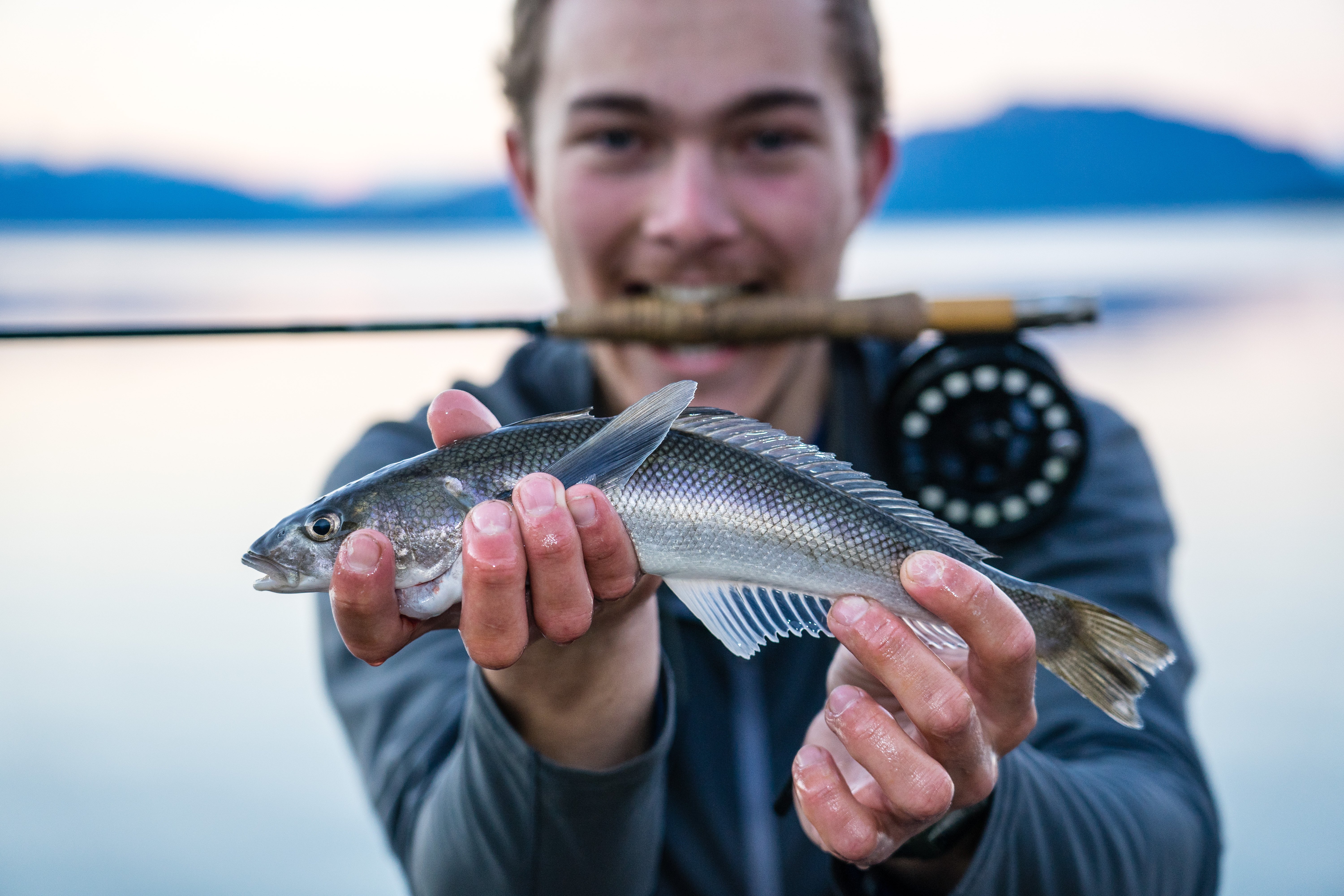 Man bites fishing rod and holds out the fish he caught with mountains and lake in the background