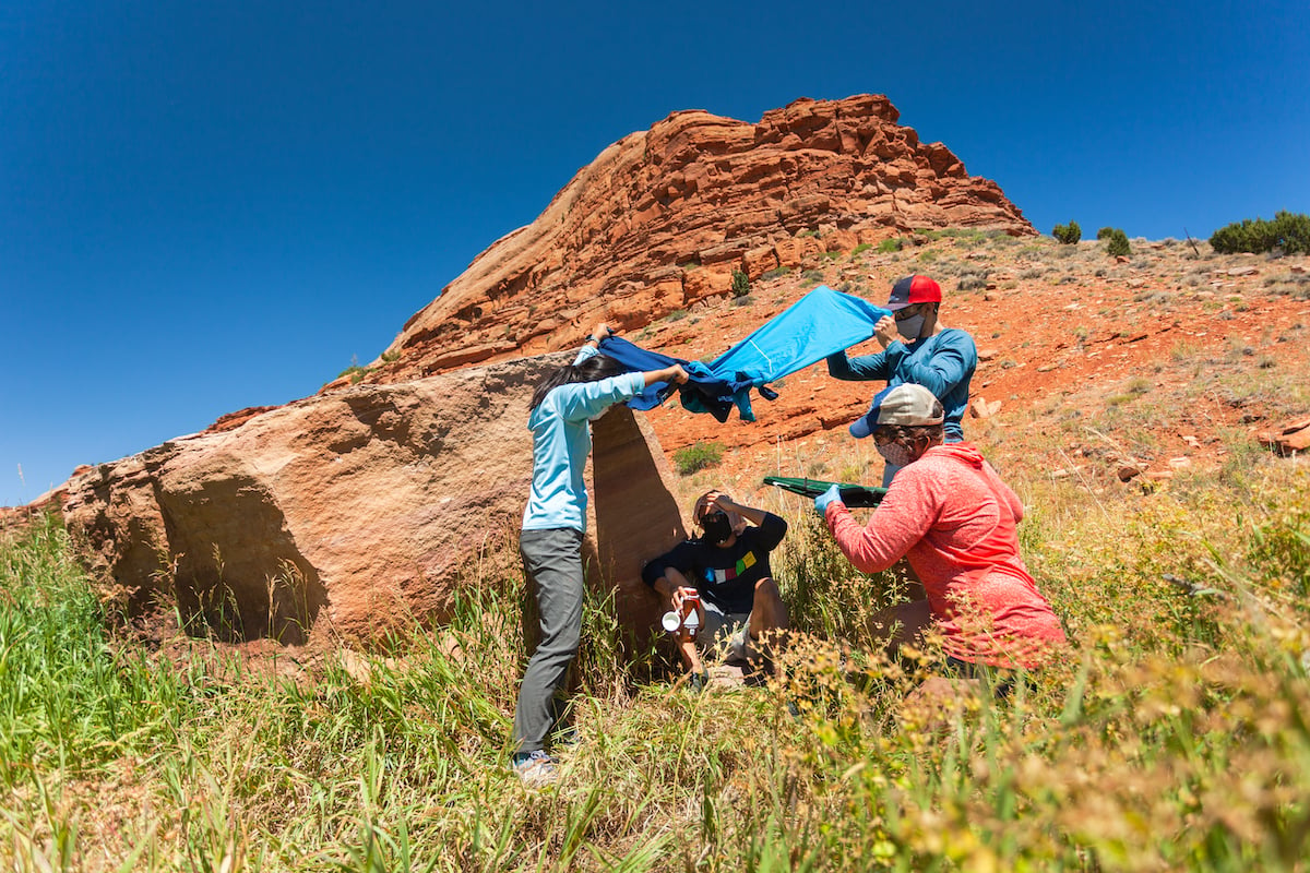 Three people improvise shade for a person practicing being a medical patient with redrock cliffs in background