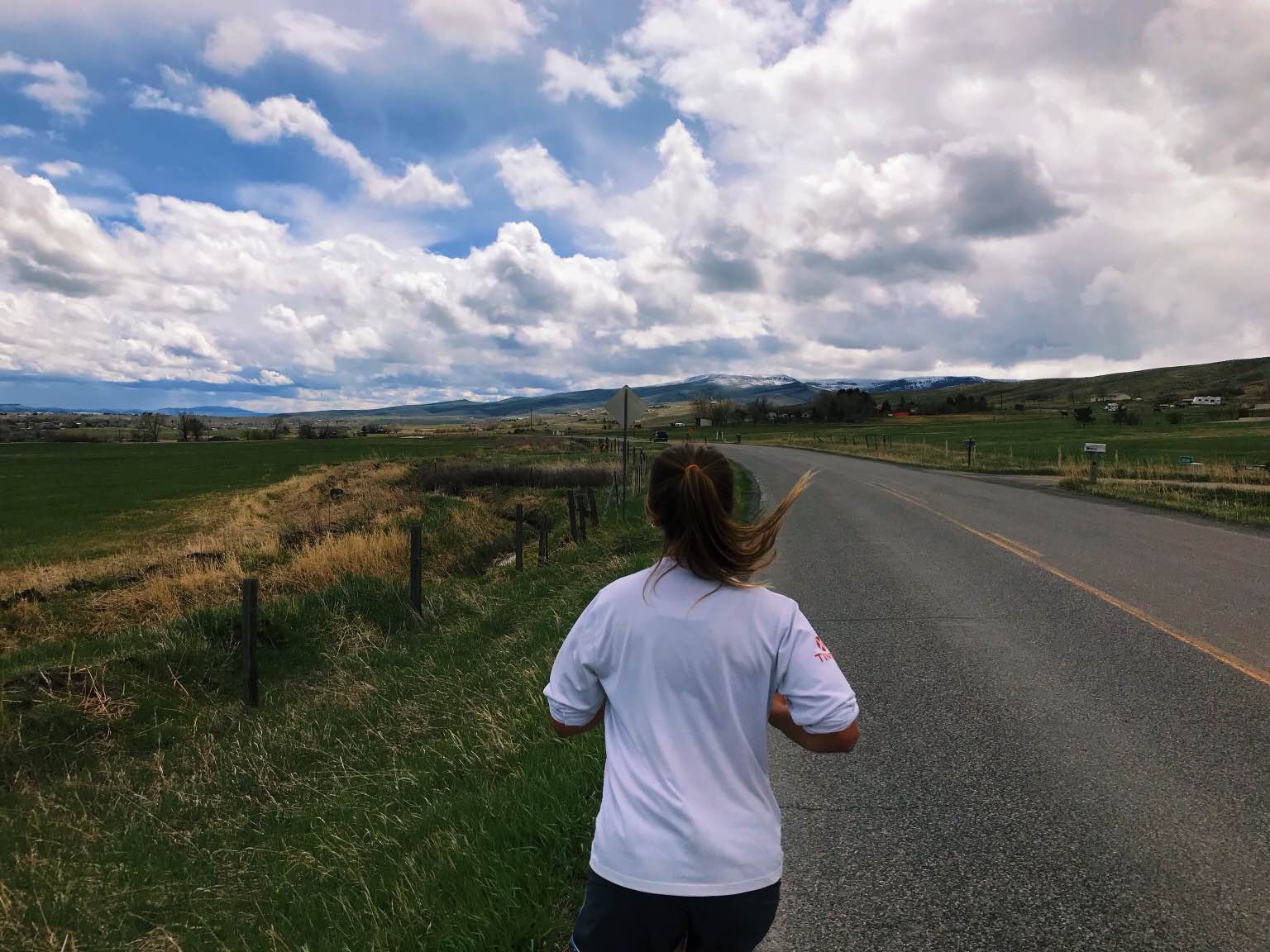 girl runs down a paved road with green fields on either side and dramatic clouds overhead