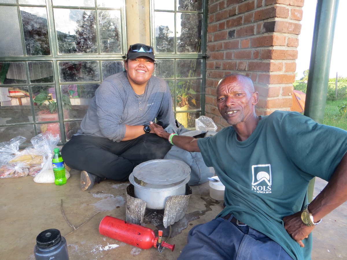 smiling NOLS student clasps hands with a Maasai hiker while making chai tea over a Whisperlite stove in Tanzania