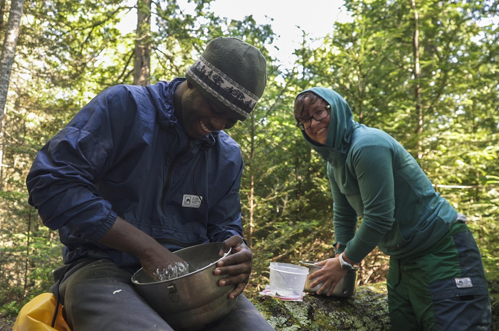 smiling NOLS student wearing windbreaker and hat makes dough in a tin bowl while another smiling participant looks on