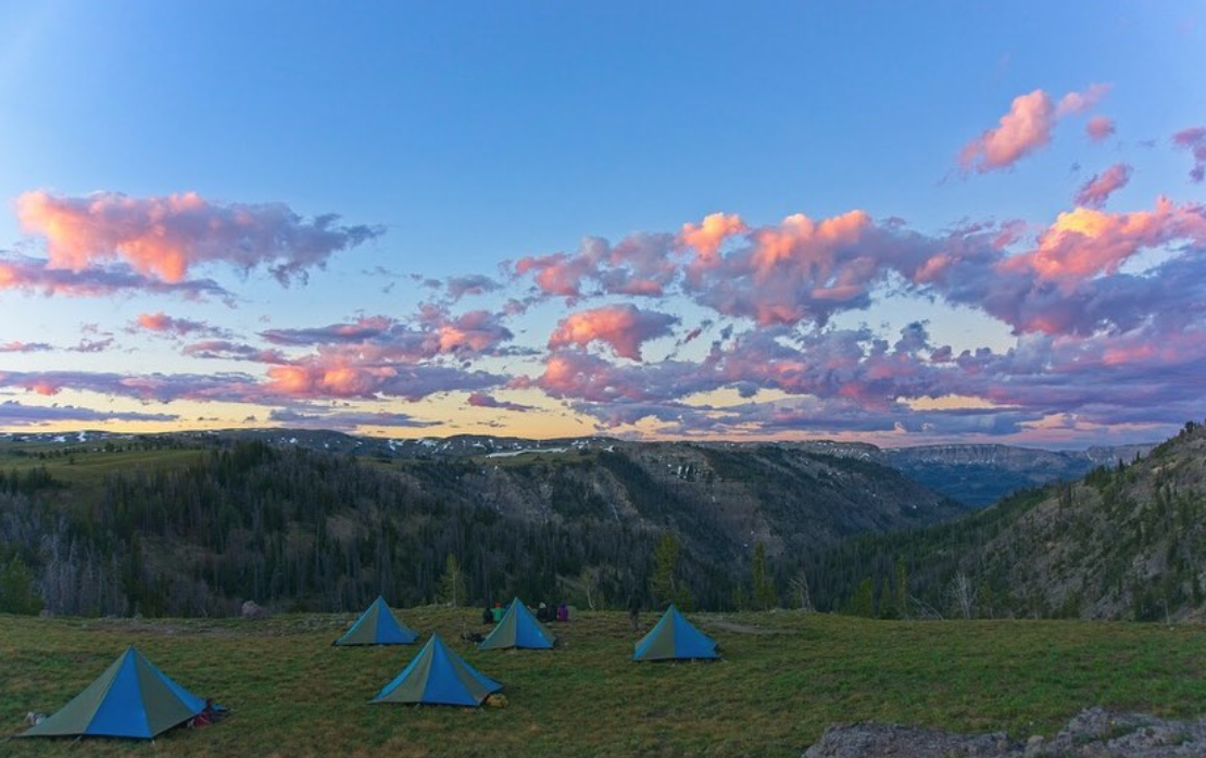 five blue and tan NOLS mega mid tents set up in a green meadow in the mountains at sunset