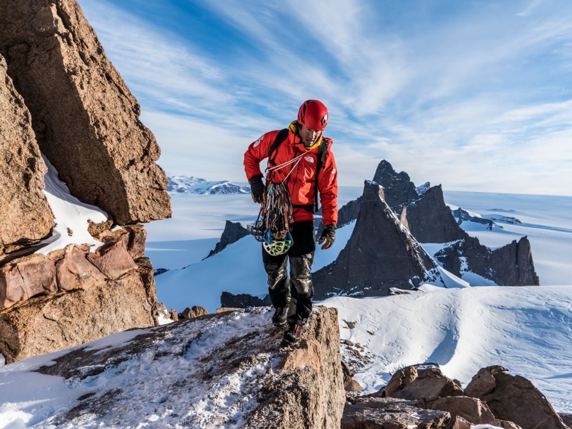 Jimmy Chin walks on a snowy rock ridge with a climbing rack of gear and a red jacket and helmet