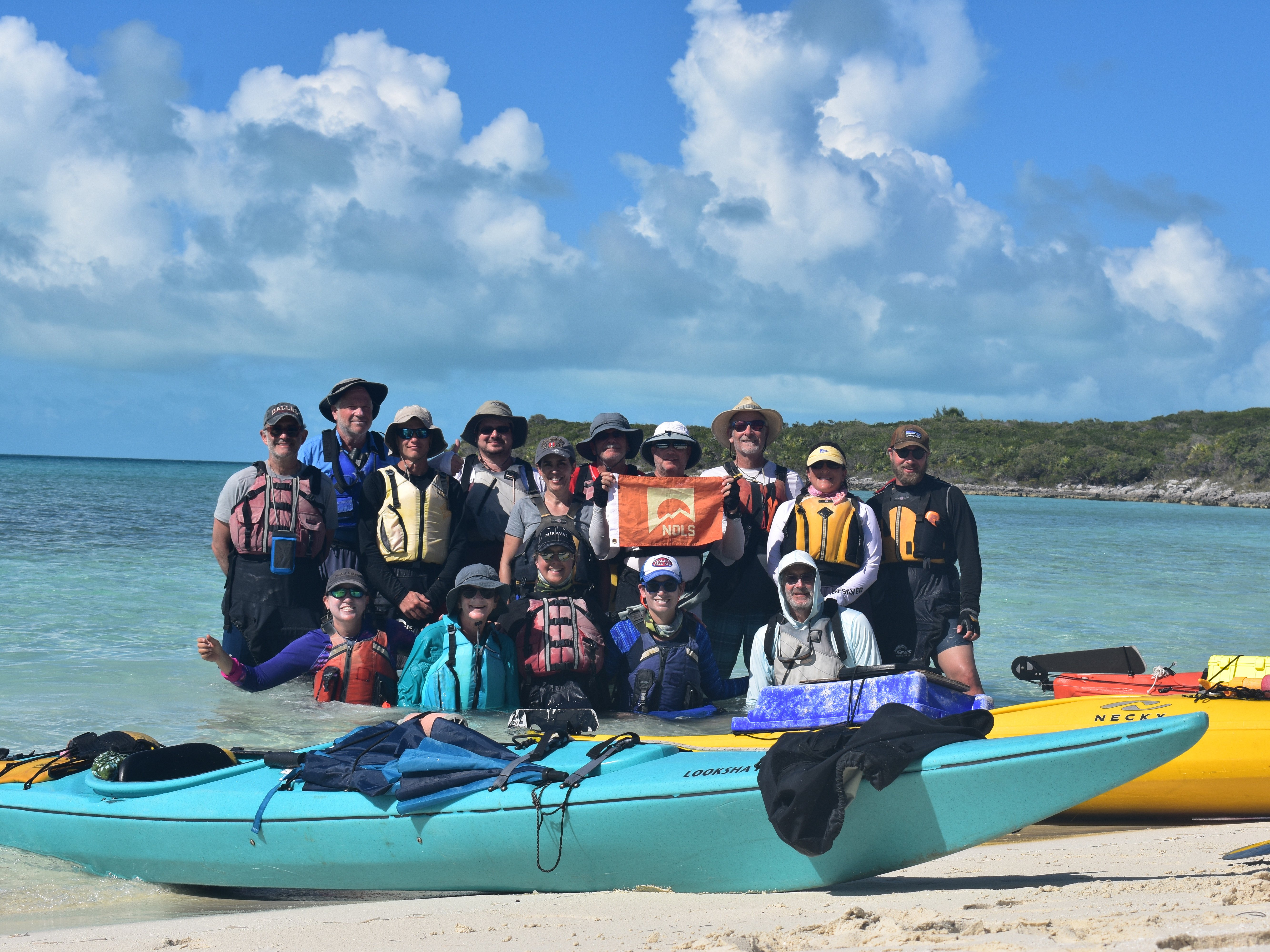 15 NOLS alumni students holding a NOLS flag while standing in the water on a beach in front of their kayaks.