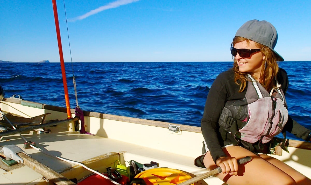 smiling woman wearing sunglasses and life preserver at the oars of a sailboat in Baja