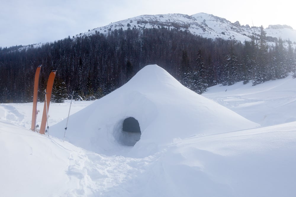 A quinzhee snow shelter stands in front of a mountain with a pair of skis outside its door