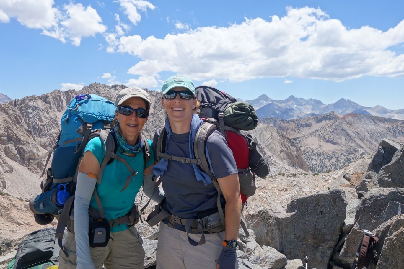 two female backpackers wearing sungasses and hats stand together high in the mountains