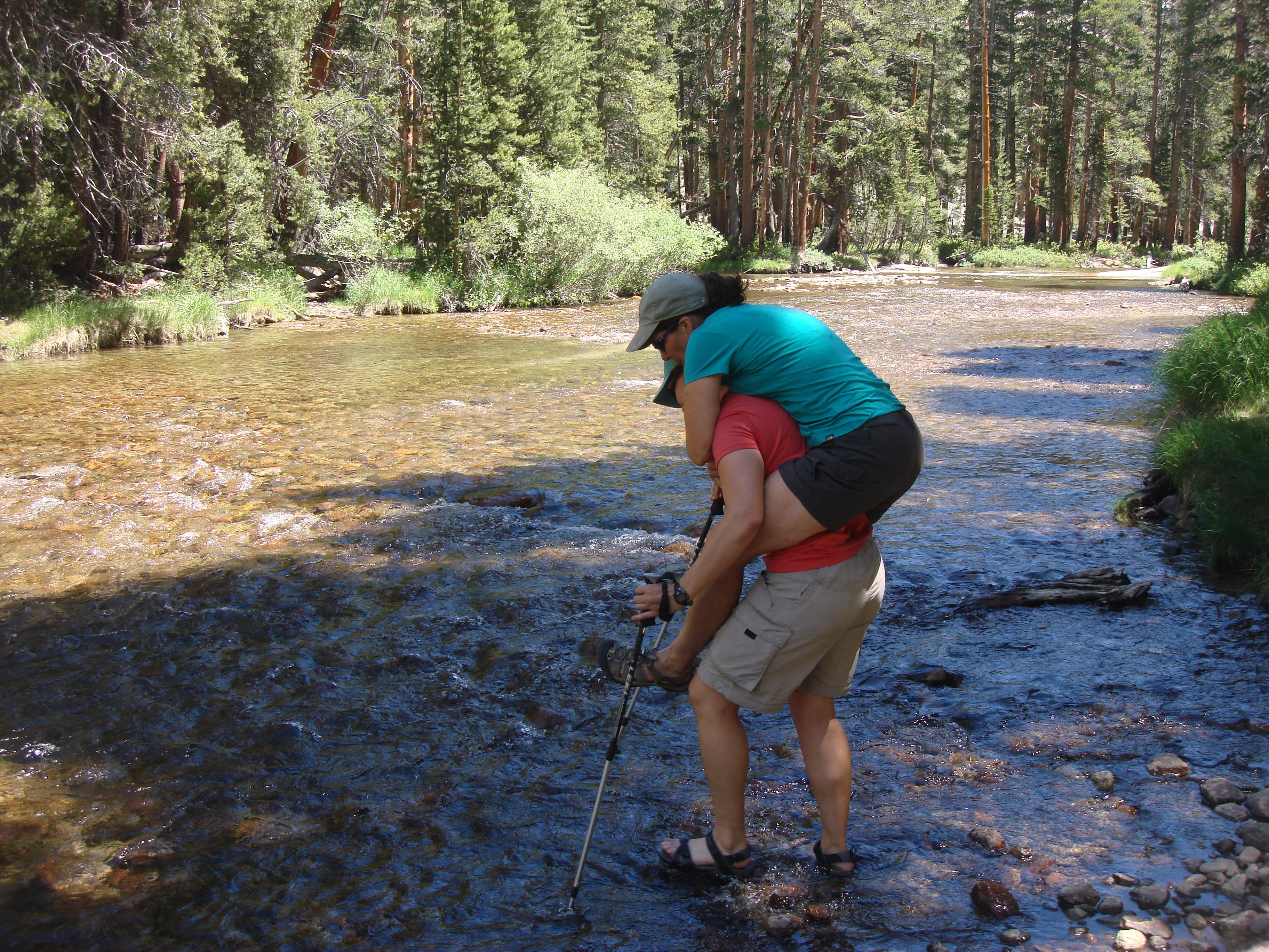 woman in a red shirt carries woman in a blue shirt across a river in summertime