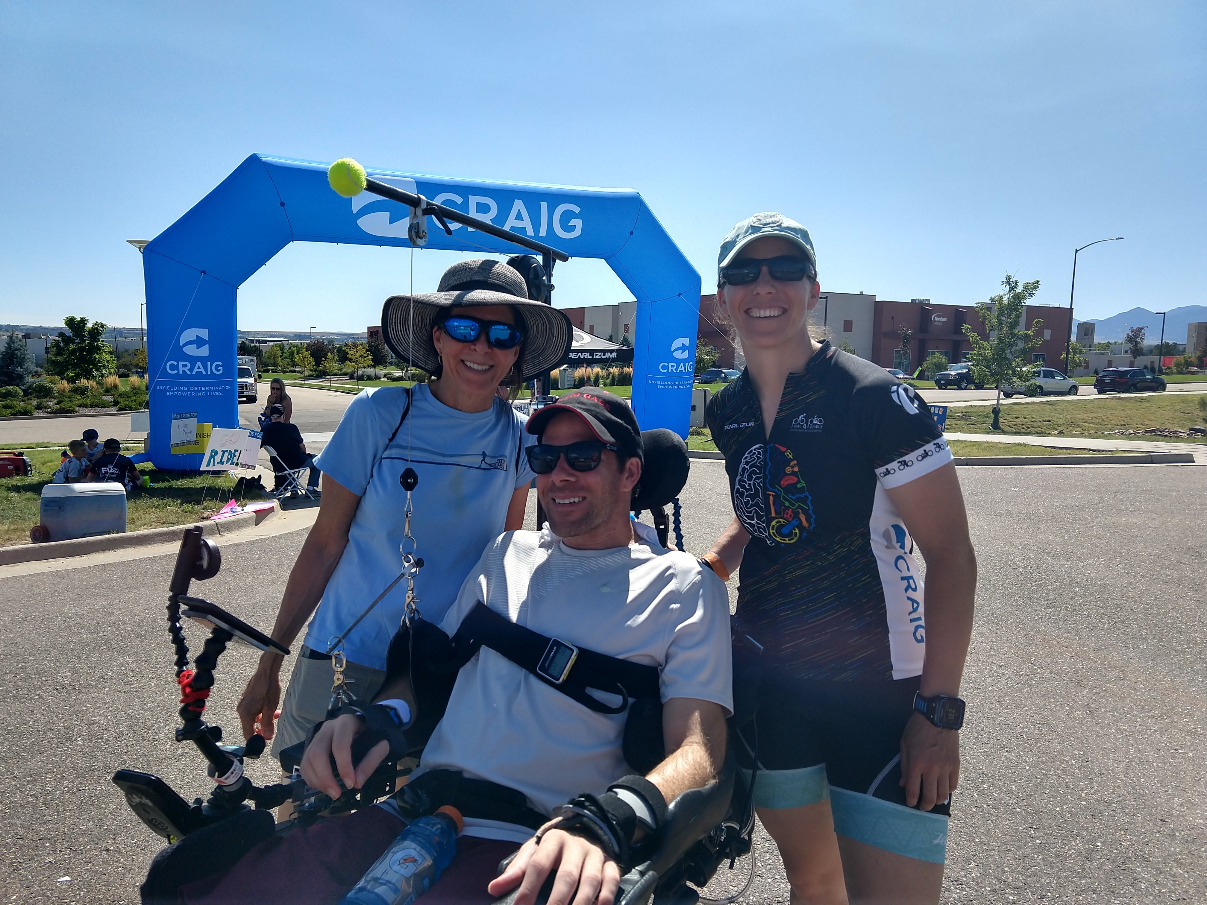 Two women, Audrey and her mom, smile in a family photo with her twin, Cory. Cory is in an electronic wheel chair. They stand in front of a bike race finish line-Audrey is in bike racing attire.