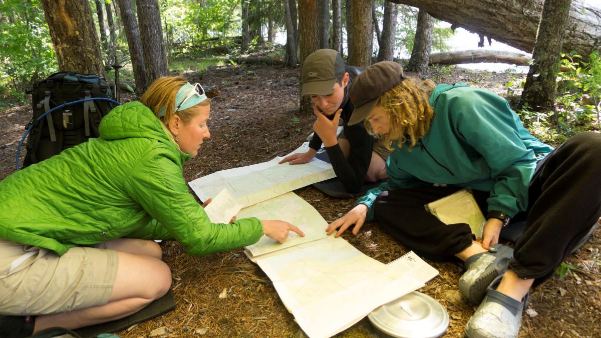 an instructor and two students study maps spread out on the ground in the woods