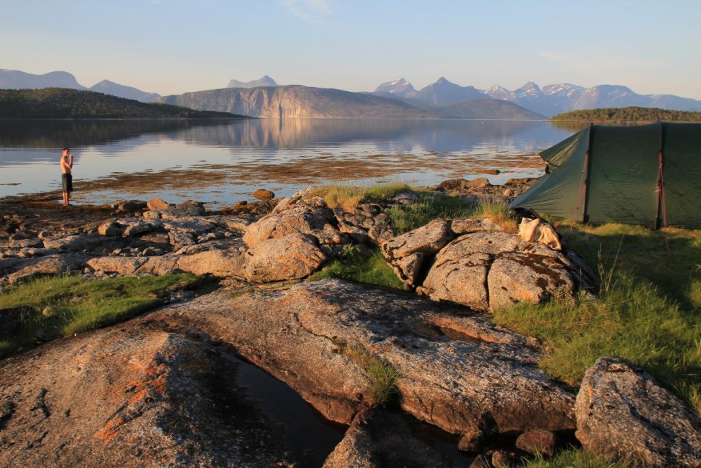 boy stands on a rocky shoreline with mountains reflected in glassy water and a green tent nearby 