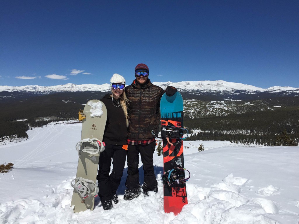 Two snowboarder smile and pose with their boards at the top of a snowy hill