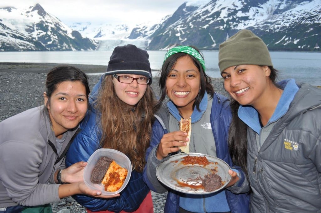 four smiling students show off their backcountry cooking skills with snowy mountains behind