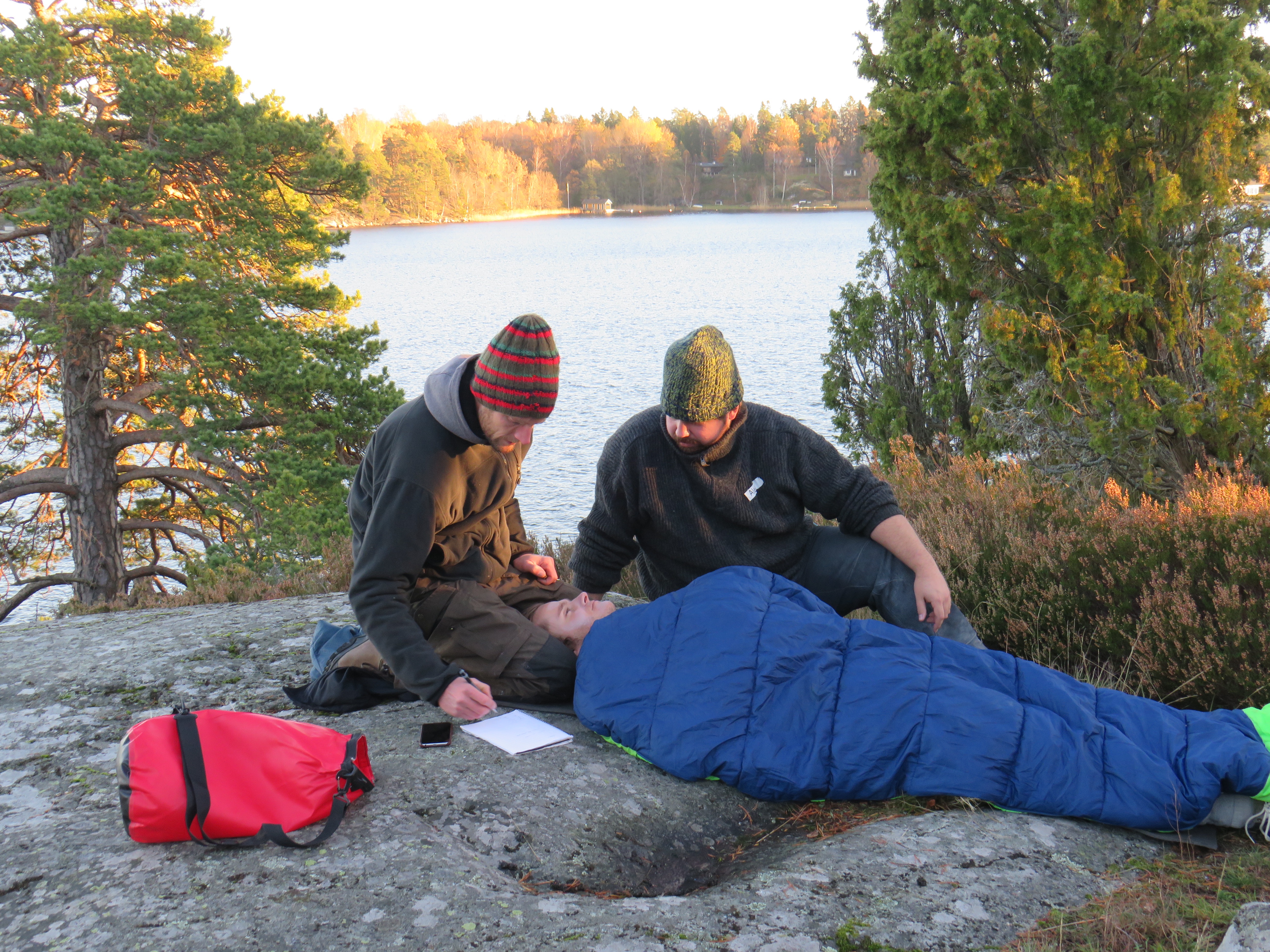 two NOLS Wilderness Medicine students practice caring for a patient lying in a sleeping back on a rock overlooking a lake