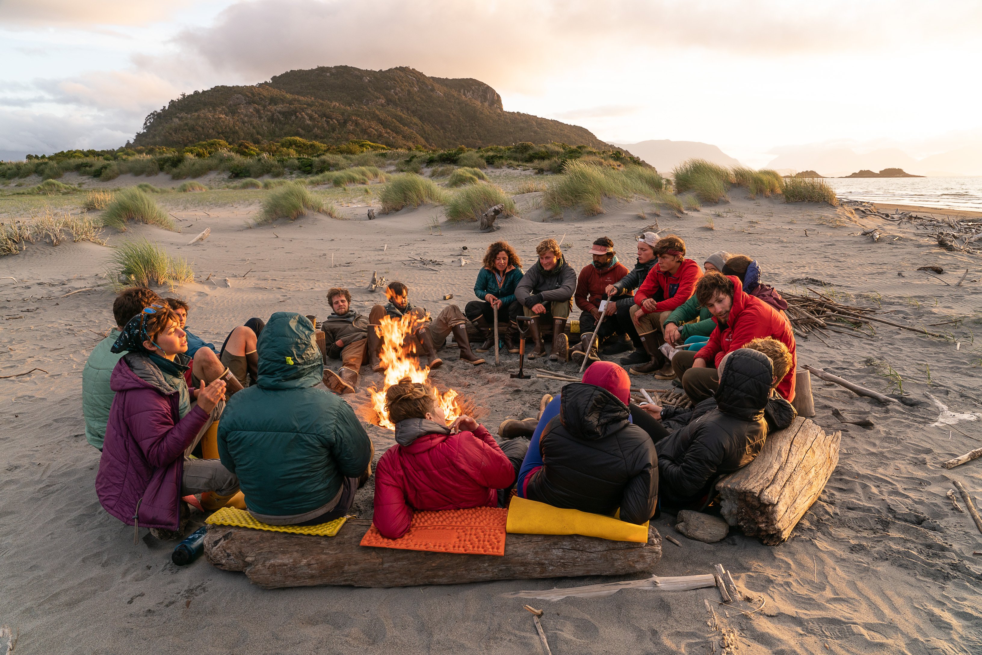 A group of people sit around a campfire on a beach. The sun is setting in the background