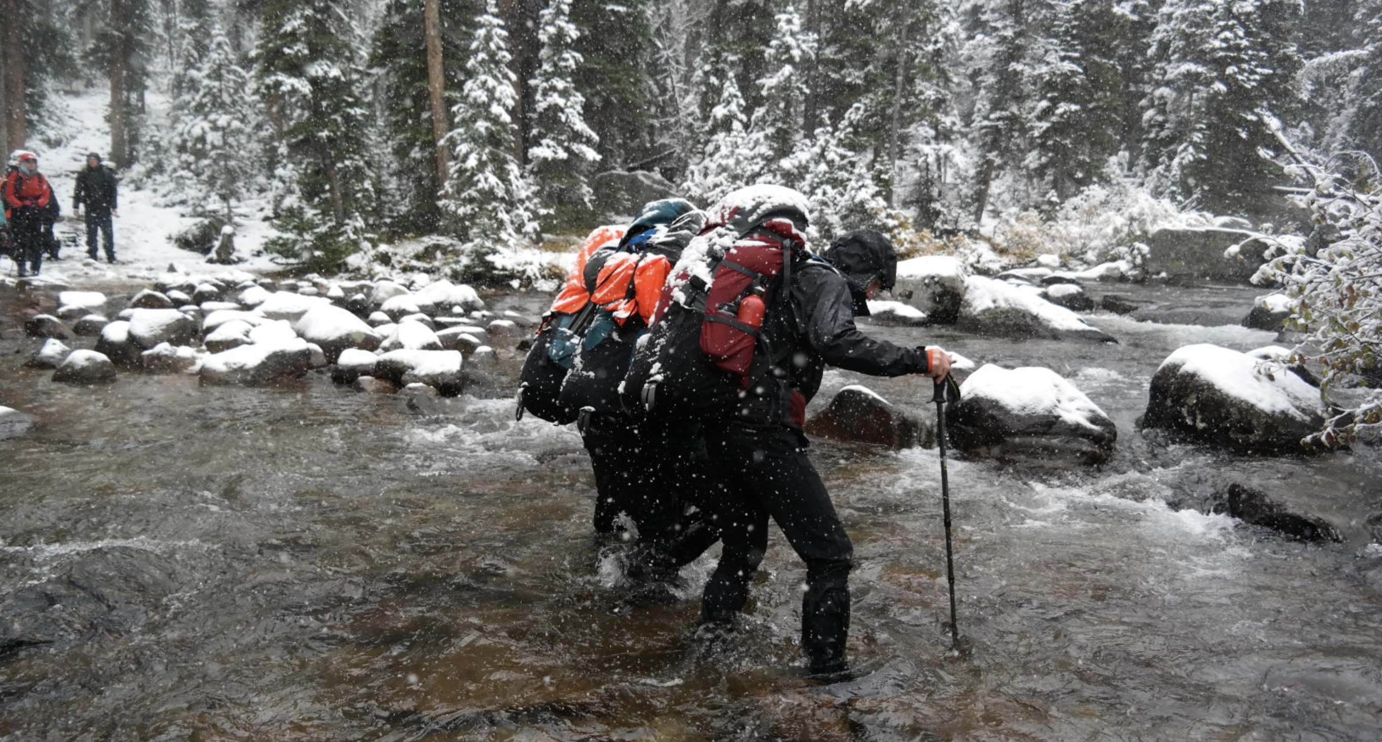 Two men with heavy backpacks cross a snowy river