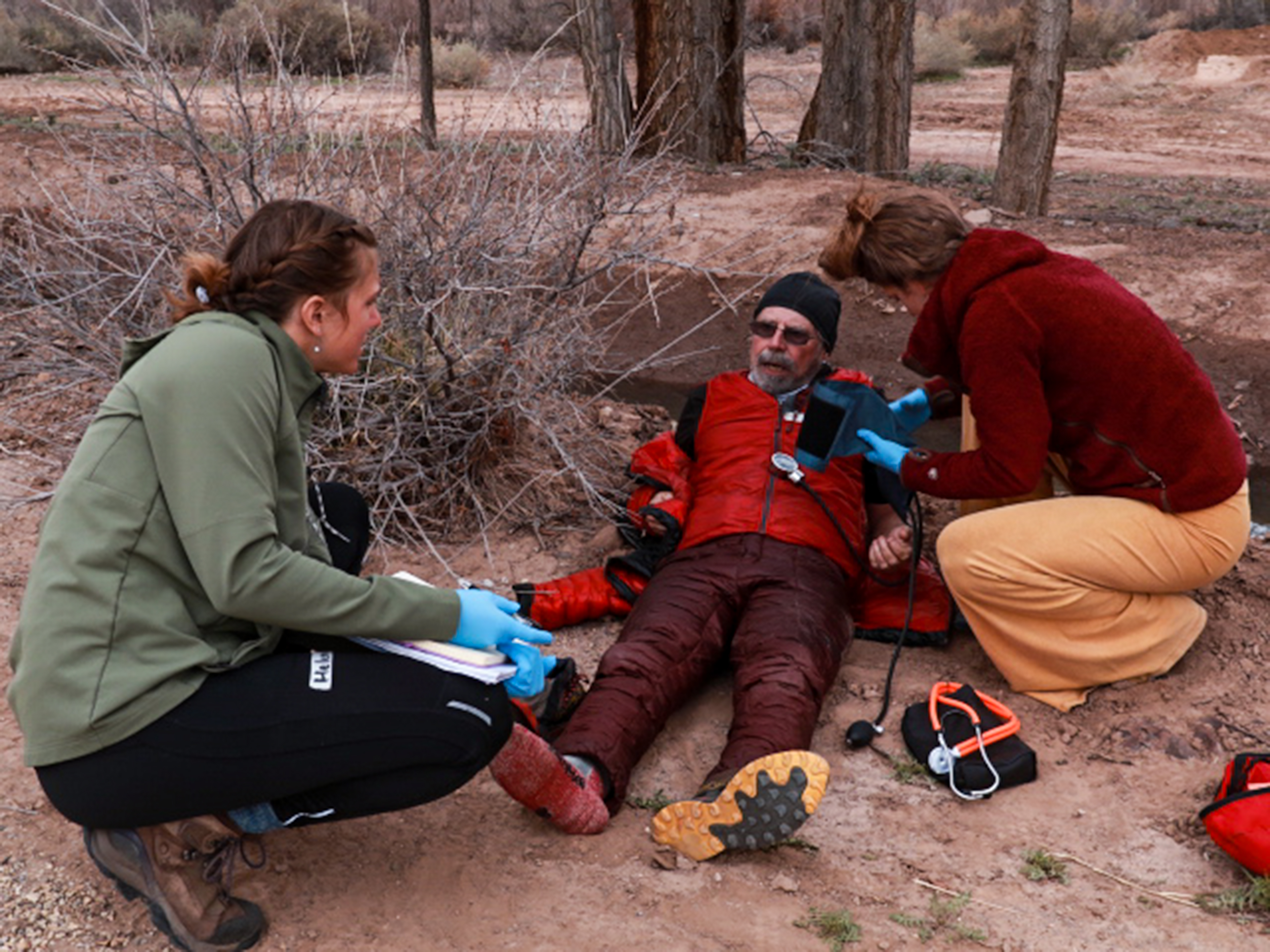 Two female rescuers kneel next to an older who is sitting on the dirt. One takes his blood pressure.